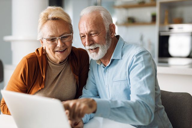 Older couple smiling and engaging with a laptop, representing seniors exploring dental health information online.