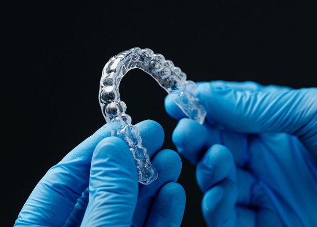 Clear aligner held by gloved hands, showcasing transparent orthodontic device designed for teeth straightening at Littleton Advanced Dentistry.