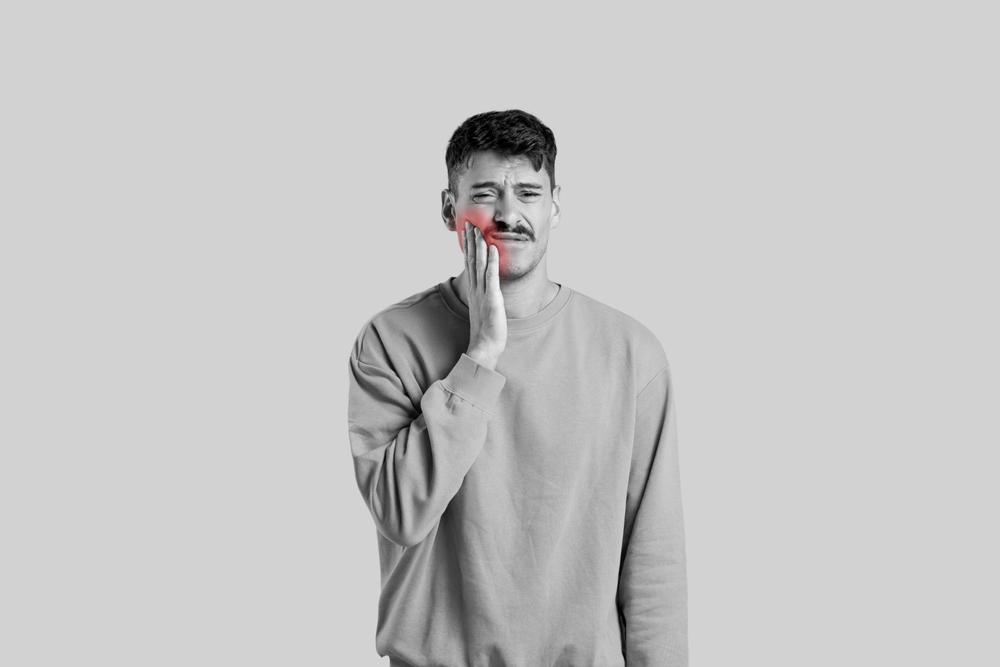 Man holding his jaw in pain, indicating a dental emergency, wearing a gray sweatshirt, against a neutral background, reflecting the urgency of dental care at Littleton Advanced Dentistry.