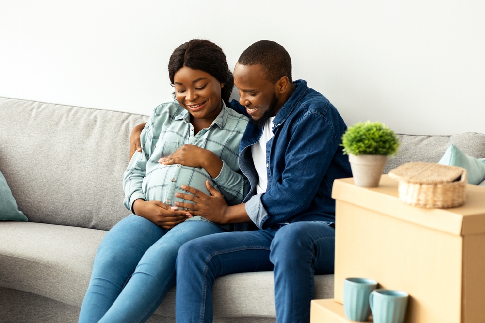 Pregnant woman sitting on a couch smiling, with her partner gently touching her belly, symbolizing prenatal care and family support during pregnancy.