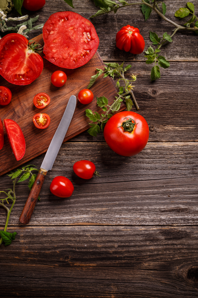 Fresh red tomatoes, sliced and whole, on a wooden cutting board with a knife, surrounded by green leaves, illustrating healthy food options for dental health.